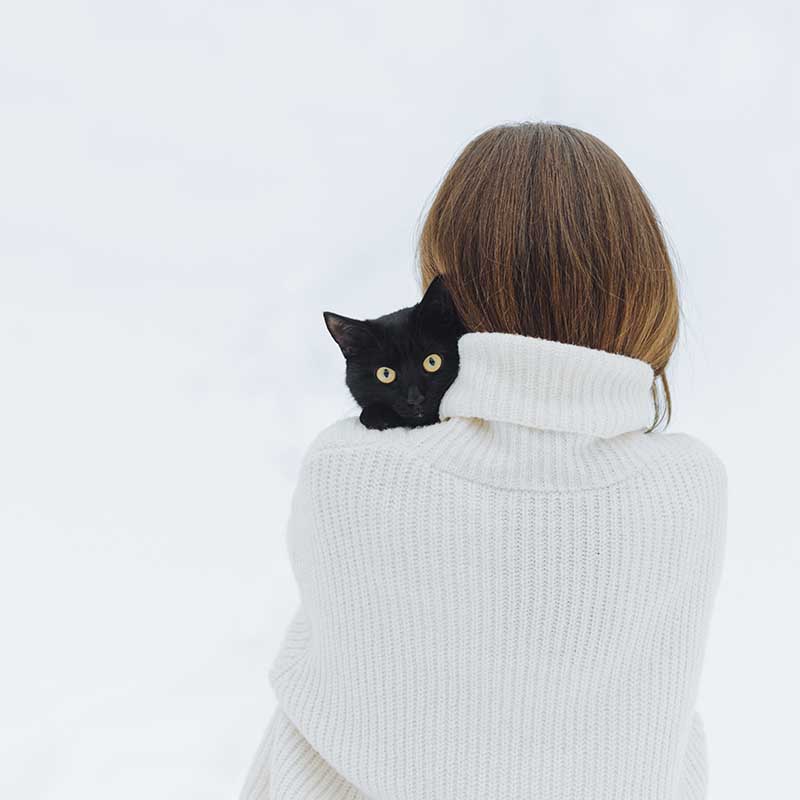 Rückansicht einer Frau im weißen Pullover mit einer schwarzen Katze, die über ihrer Schulter in die Kamera schaut.