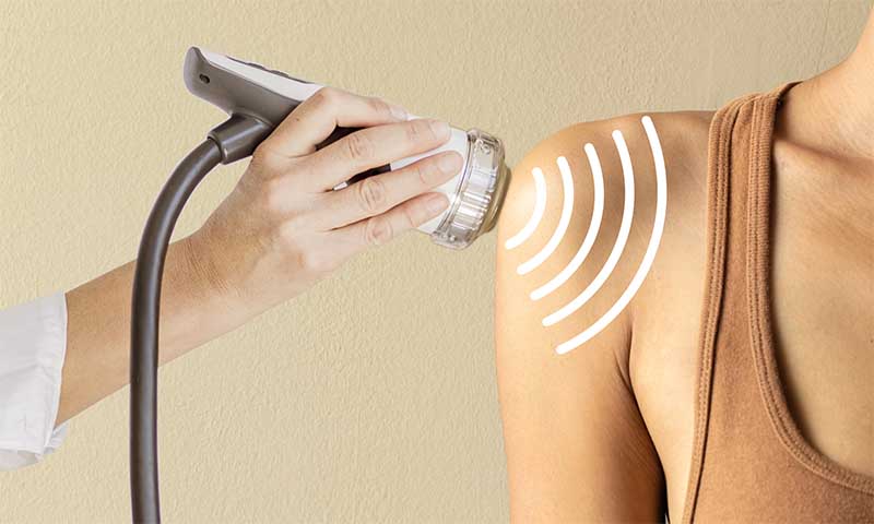 Ein Orthopäde behandelt Schulter einer Frau mit Stoßwellentherapie-Gerät